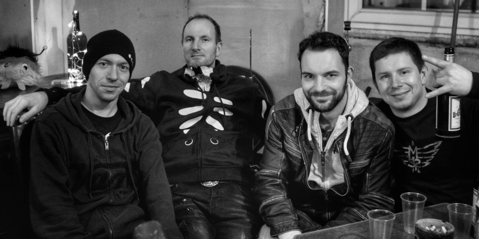 Ein Bild von Tobi, Josch, Alexander und Mart aus dem Backstagebereich eines Konzerts. Sie schauen freundlich und prosten dem Bildbetrachter zu.