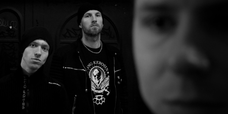 Ein Bandfoto der drei Bandmitglieder, bei denen man Tobi und Josch deutlich im Hintergrund sieht und verschwommen im Vordergrund das Gesicht von Mart.